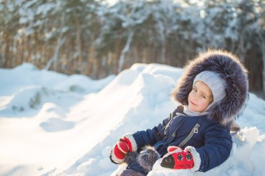 Kış elbisesi içinde, bir kış ormanının arka planında poz veren sevimli, güler yüzlü bir kızın portresi. Kışın açık hava aktiviteleri kavramı