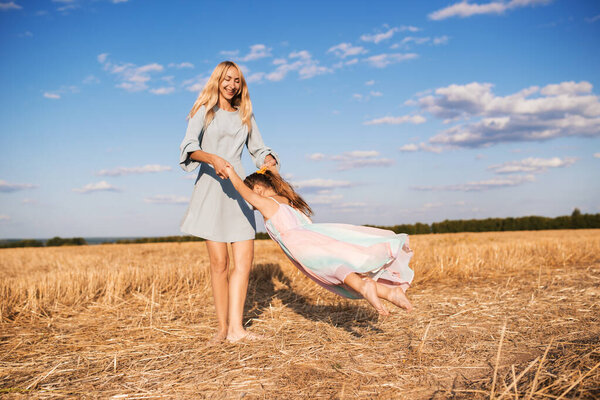 Cheerful young mother in a blue dress is spinning with her little beautiful daughter standing on the field barefoot on a warm sunny summer day. Child care concept