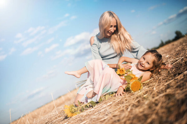 Charming young mother is having fun with her little daughter sitting on a field with sunflowers on a warm sunny summer day. Weekend vacation concept with family