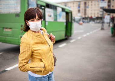Beyaz maskeli, orta yaşlı bir kadının portresi ve otobüs durağında toplu taşıma için poz veren sıradan bir kadın. Salgın ve koronovirüs koruma koşulları kavramı