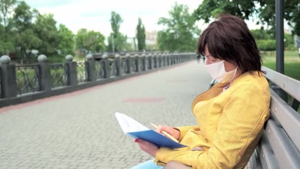 Vrouw met een beschermend masker zit op een bankje in het park en maakt aantekeningen in haar notitieboekje. — Stockvideo