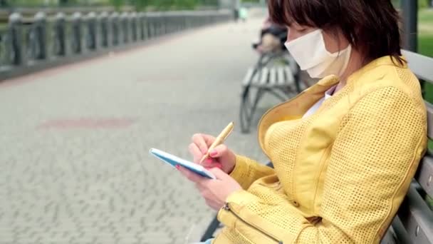 Donna in maschera protettiva si siede su una panchina del parco e prende appunti nel suo quaderno . — Video Stock