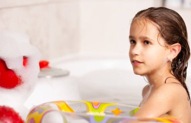 Komik küçük kız küvette şişme bir can simidiyle ve suda köpükle banyo yapıyor. Çocuklar için hijyen ve eğlence kavramı. Reklam alanı
