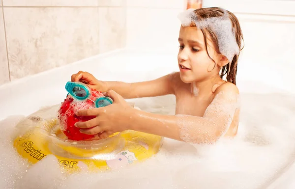 Funny little girl bathes in a bath with foam and plays life saving ball and swimming goggles. Children leisure at home concept. Copyspace