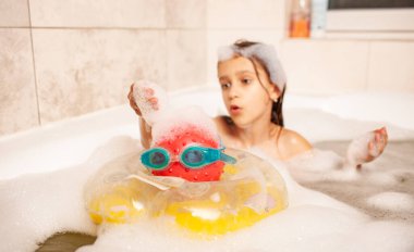 Komik küçük kız köpükle banyo yapıyor ve hayat kurtaran top ve yüzme gözlüğü oynuyor. Çocuklar ev kavramında boş vakit geçirirler. Kopyalama alanı