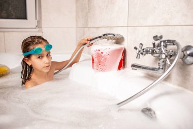 Komik küçük kız küvette banyo yapıyor ve duştaki beyaz köpüğe su döküyor. Karantina sırasında evde suyla yapılan çocuk oyunları kavramı