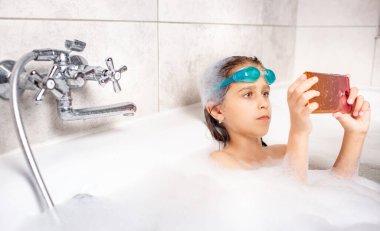 Yüzme gözlüklü, odaklanmış beyaz bir kız evde köpüklü bir küvette banyo yaparken akıllı telefon kullanarak selfie çekiyor. Corovirüs sırasındaki çocuk ve aletler kavramı