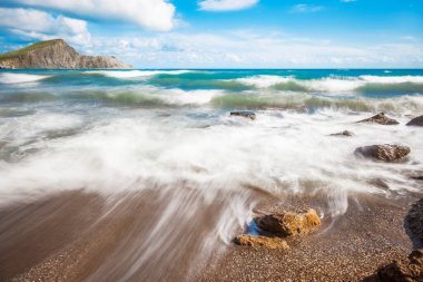 Arkasında dağlar olan kumsal. Dağlar çimlerle kaplıdır ve denizden dik kayalıklara sahiptir. Gökyüzü bulutlu