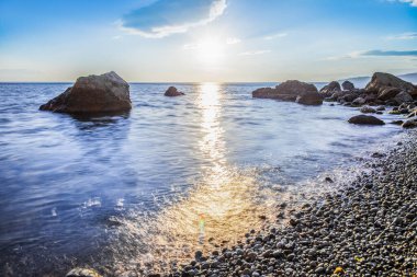 Okyanustaki taşlar gün batımına karşı, hangi sisin üzerinde. Gökyüzü bulutlarla dolu. Ufkun ötesinde dağlar var.