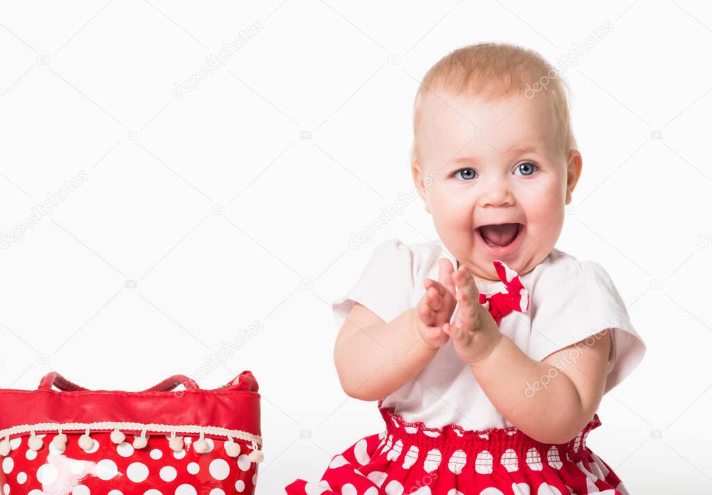 Front view of cute child with wide opened mouth looking aside and applauding. Joyful kid in stylish dress sitting next to polka-dot bag. Isolated on white studio background. Concept of baby cuteness.
