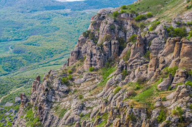 Ön planda yosunlarla kaplı dev bir taş dağın yamacındaki ağaçlar, vadideki yeşil çimenler.