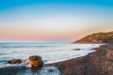 Deniz kıyısında gün batımı, kayalık plaj ve mavi gökyüzü. Arka planda yeşil bitkilerle kaplı küçük tepeler