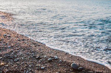 Deniz kıyısı ve kayalık sahil, beyaz bulutlu mavi gökyüzü, dağlar ve arka planda küçük bir köy.