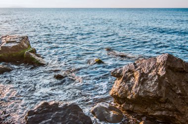 Deniz kıyısında gün batımı, kayalık plaj ve mavi gökyüzü. Arka planda yeşil bitkilerle kaplı küçük tepeler
