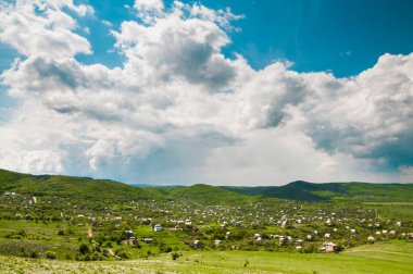 Dağların arasında küçük bir köyün fotoğrafı. Etrafta bir sürü yeşil bitki, bulutlu gökyüzü