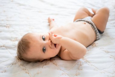 Evdeki beyaz yatakta pantolonunu giymiş tatlı küçük bebek kameraya bakıyor. Beyaz tenli beyaz bir bebeğin parmaklarını ağzında tutmasının en iyi görüntüsü. Çocukluk kavramı, bebekler.