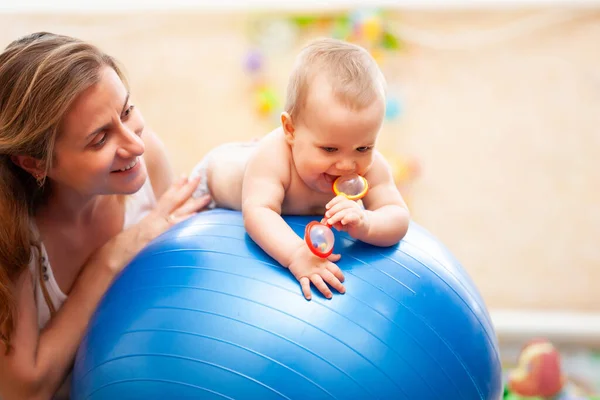 可爱的幼儿有选择的焦点躺在蓝色的大健身球上 拿着红色的玩具 赤身裸体的高加索孩子为婴儿练体操的镜头 快乐的母亲在背后帮忙 — 图库照片