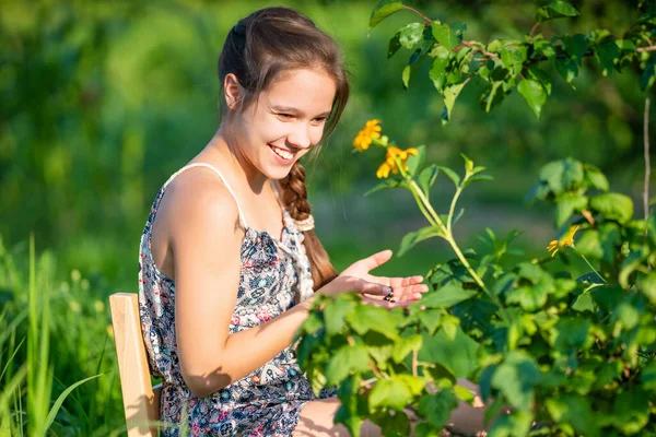 Kahverengi örgülü beyaz kadın gencin seçici odağı çalılık ve çiçeklerin yanındaki ahşap sandalyede oturuyor. Kırsal kesimde hasat toplayan çiftçi kız manzarası.
