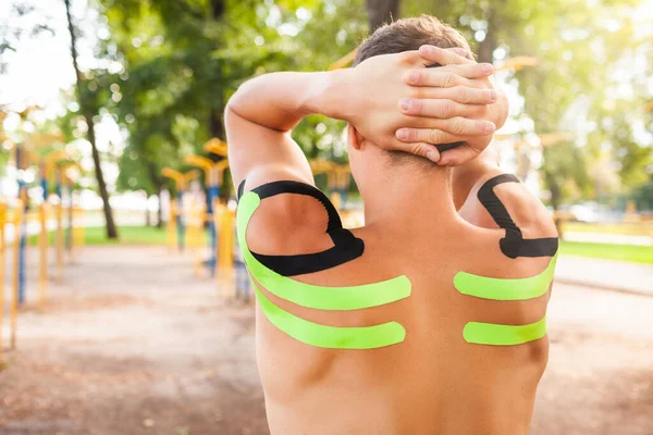 Spor sahasında poz veren çıplak kaslı omuzlarda siyah ve yeşil kineziyolojik bantları olan tanınmayan beyaz profesyonel vücut geliştirici. Kolları dışarıda uzanan boynunun arkasında bir adam..