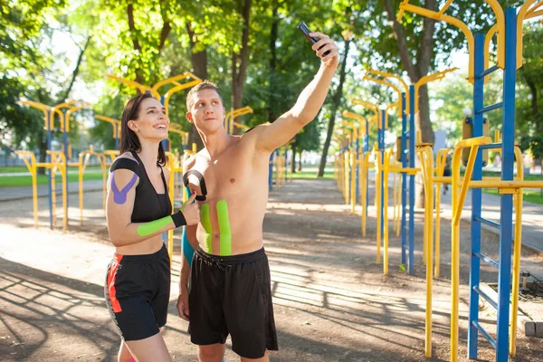 Kineziyoloji elastik bantlı profesyonel beyaz atletler, yakışıklı erkek ve esmer kadın, spor sahasında poz veriyor, dinleniyor ve akıllı telefon kullanarak selfie çekiyor..