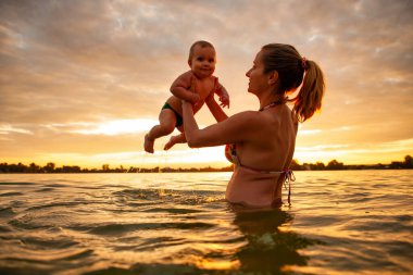 Sıcak deniz suyunun üzerinde gülümseyen küçük tatlı bir çocuğu tutan mutlu annenin yan görüntüsü. Beyaz bir aile, parlak turuncu günbatımında yaz tatilinin tadını çıkarıyor. Aile kavramı.
