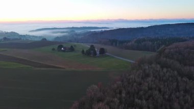 Geniş ve dolambaçlı otoyollar, koyu yoğun çam ormanları, çimenli yeşil çayırlar ve güneşli akşam ışığında beyaz sisle kaplanmış küçük bir köy ile yuvarlanan bir vadiden geçer. Video 4k UHD eş zamanlı