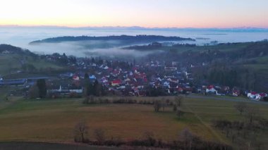 Geniş ve dolambaçlı otoyollar, koyu yoğun çam ormanları, çimenli yeşil çayırlar ve güneşli akşam ışığında beyaz sisle kaplanmış küçük bir köy ile yuvarlanan bir vadiden geçer. Video 4k UHD eş zamanlı