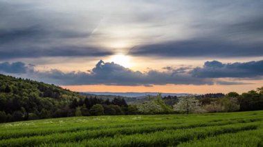 Gri bulutlu bir gökyüzünde parlak güneşli bir akşam günbatımı beyaz meyve bahçeleri, çimenli çayırları ve karanlık yoğun ormanlarıyla tepelik geniş bir kırsal vadiyi aydınlatır. Video 4k UHD zaman ayarlı