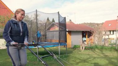 Koyu renk yelek giyen kızıl saçlı, çalışkan bir yetişkin kız arka bahçesinde güçlü, modern bir çim biçme makinesiyle taze bahar yeşili çimenleri biçiyor ve oğlu trambolinde zıplıyor. 4k video yavaş çekim UHD