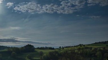 Mavi-gri bulutlu gökyüzünde parlak bir yaz akşamı güneşi tepelerin yeşil çayır yamaçları ve karanlık bahar ormanları ile devasa bir yaban vadisini aydınlatır. Video 4k UHD zaman ayarlı