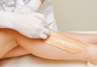 Cosmetologist beautician waxing female legs in the spa center be clipart