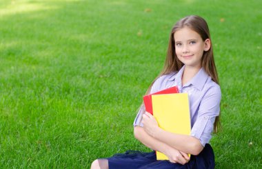 Okula dönelim. Eğitim konsepti. Çimlerin üzerinde oturan sevimli gülümseyen kız öğrenci. Kitapları tutan mutlu küçük kız çocuğu