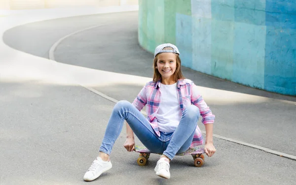 スケートボードに座ってキャップでかわいい女の子の子供を笑顔 屋外で夏の日にペニーボードでプレティーン — ストック写真