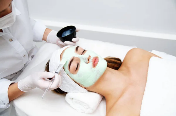 Beautiful young woman with facial mask in beauty salon. Girl getting beauty treatment facial care by beautician at spa salon.