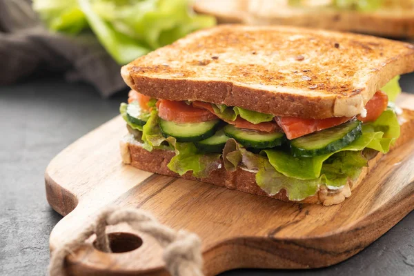 Sandwich with white bread toasts, red fish salmon, fresh green leaves of salad and sliced cucumber on a wooden board.