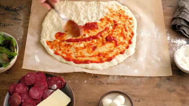 Pemandangan teratas, tangan wanita mengolesi adonan dengan saus, menyiapkan pizza Italia dengan saus, tomat, bumbu, sosis pepperoni, bayam, sayuran, keju mozzarella. Dough untuk roti. Pizza pepperoni. — Stok Video