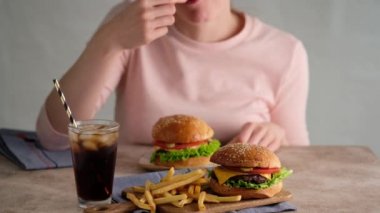 Fast food yiyen bir kadın. Hamburger, patates kızartması, kola. Bir kadın çörek, pirzola, marul, salatalık, domates, soğan ve sostan hamburger yaptı. Güzel arka plan. Yavaş çekim.