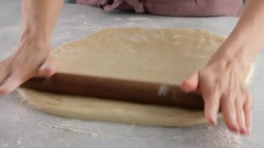 Hamuru merdaneyle yuvarlayıp hamuru şekillendiriyor. Masada un içinde hamur yoğuran kadın eller. Kapatın. Tarçınlı çörek üretimi. Fırın ürünleri.
