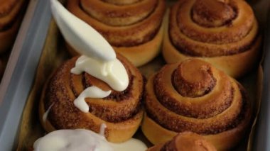 Taze pişmiş tarçınlı çöreklere ya da Cinnabon 'a krema dök. Tarçınlı ruloların üzerine tatlı krem peynirli krema dökülüyor. Tarçınlı çörek üretimi. Fırın ürünleri. Aperatif tarçınlı çörekler