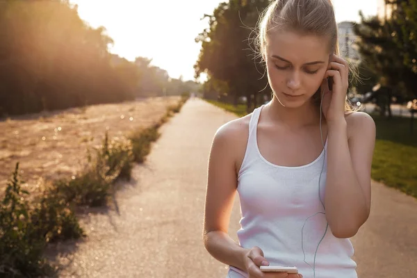 The girl walking outdoor on the road and listen to music in earphones. Woman with blonde hair and in sport clothes looking on her smartphone