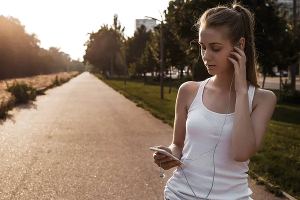 Young woman walking outdoor on the road and listen to music in earphones. The girl with blonde hair and in sport clothes looking on her smartphone.