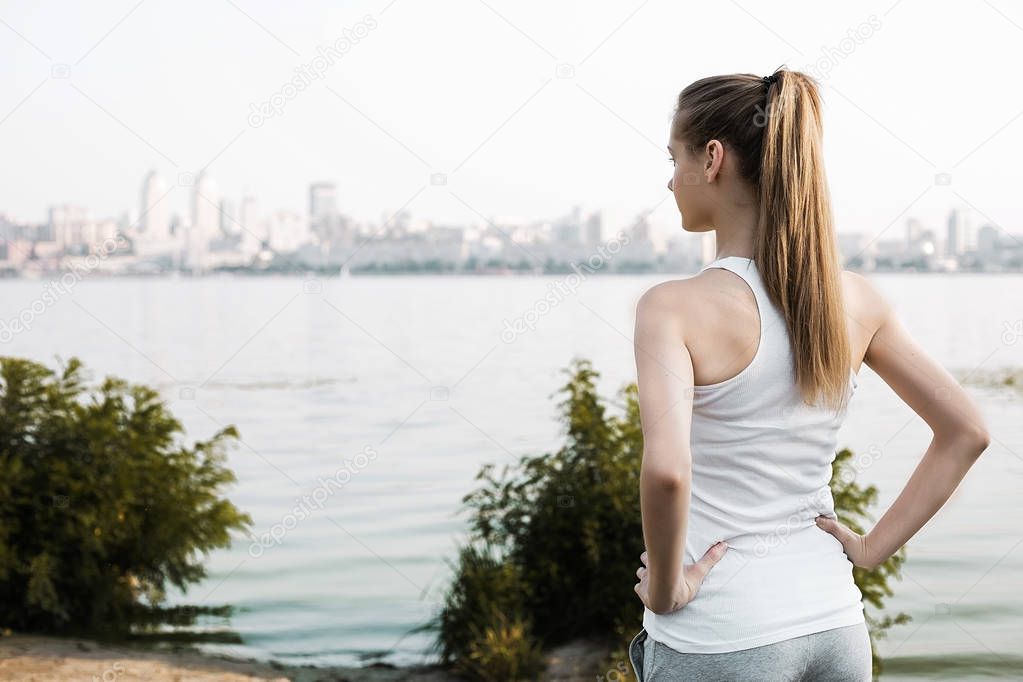 Sportwoman standing by back to camera and looking on city behind the river