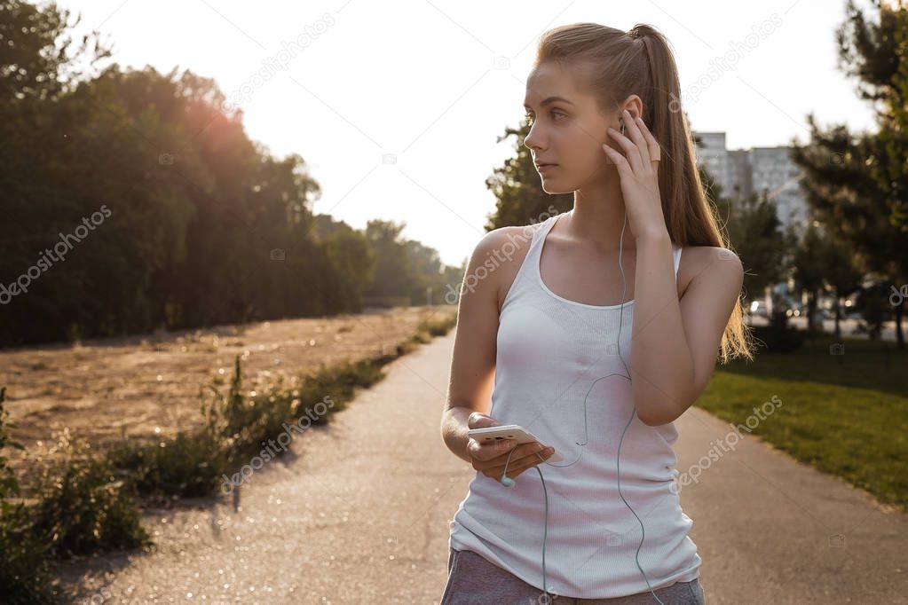 Pretty young model in sport clothes standing on the road and looking sideways. The girl listen to music in earphones