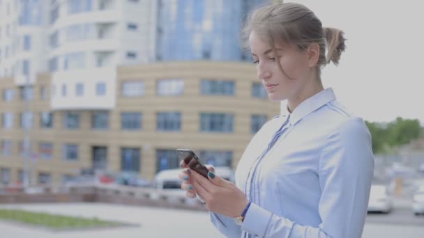 年轻的女商人用她的智能手机上网冲浪 站在街上的女孩在城市背景上 — 图库视频影像