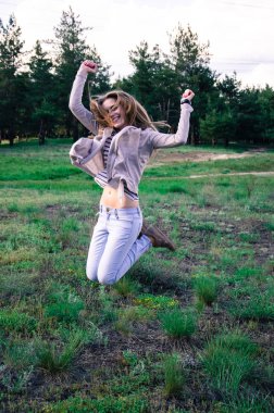 Çayırda zıplayan neşeli genç bir kadın. Sarı saçlı kız eğleniyor. Beyaz kot, ceket ve çizgili tişört giymiş bir kadın. Açık hava..