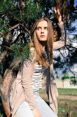 Ağaç üzerinde oturan ve kameraya poz güzel ve seksi modeli. Yana doğru seyir kız. Beyaz kot pantolon, crem ceket ve çizgili tişört giyen genç kadın. Açık. Orta atış.