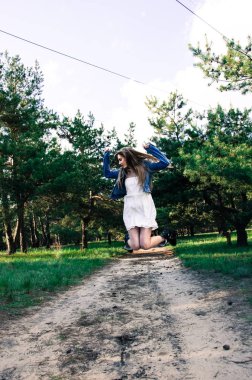 Sevimli ve Genç ve güzel sarışın kız beyaz elbise, kot ceket ve siyah çizmeler orman kum de yol atlama. Kırsal kız iyi eğlenceler, hop ve kafasını yana doğru çevirdi. Portre. Tam vuruş. Açık.