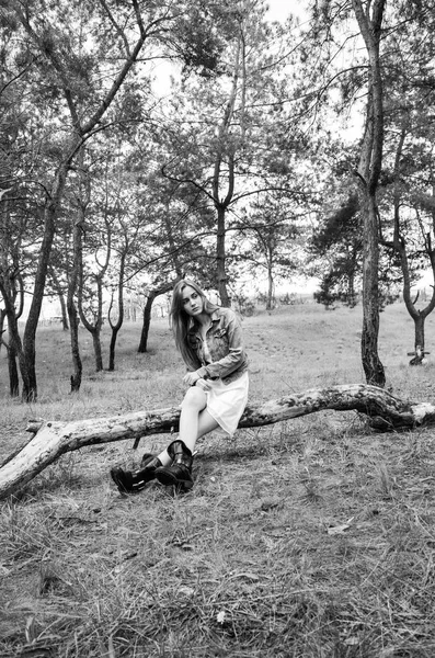 性感模特坐在木质原木上 看着带着沉思的相机 穿着白色连衣裙 牛仔裤夹克和黑色靴子的金发女郎 时髦的人摆在相机上 黑白相间 — 图库照片
