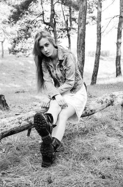 性感模特坐在木质原木上 看着带着沉思的相机 穿着白色连衣裙 牛仔裤夹克和黑色靴子的金发女郎 时髦的人摆在相机上 — 图库照片
