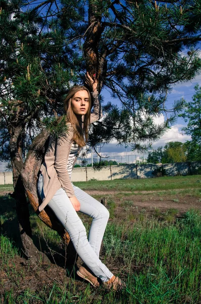 Pretty and sexy model sitting on the tree and posing on the camera. The girl looking sideways. Young woman wearing white jeans, crem jacket and striped T-shirt. Outdoor. Full shot.